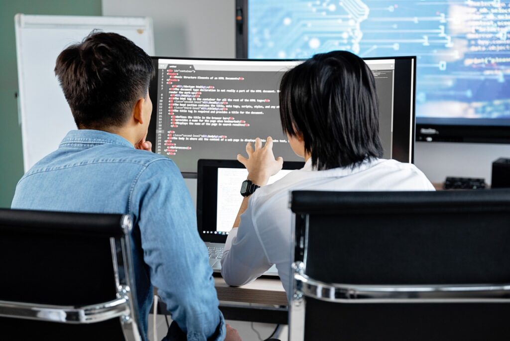 A imagem exibe duas pessoas sentadas de costas para a câmera, concentradas em uma tela de computador curvada que apresenta linhas de código de programação. O indivíduo à esquerda utiliza uma camisa jeans azul, enquanto o da direita veste uma camisa social branca. Ambos estão em cadeiras de escritório pretas diante de uma mesa de madeira. No fundo, há um quadro branco e outra tela que exibe gráficos digitais. Remete à contratação de profissionais de tecnologia.